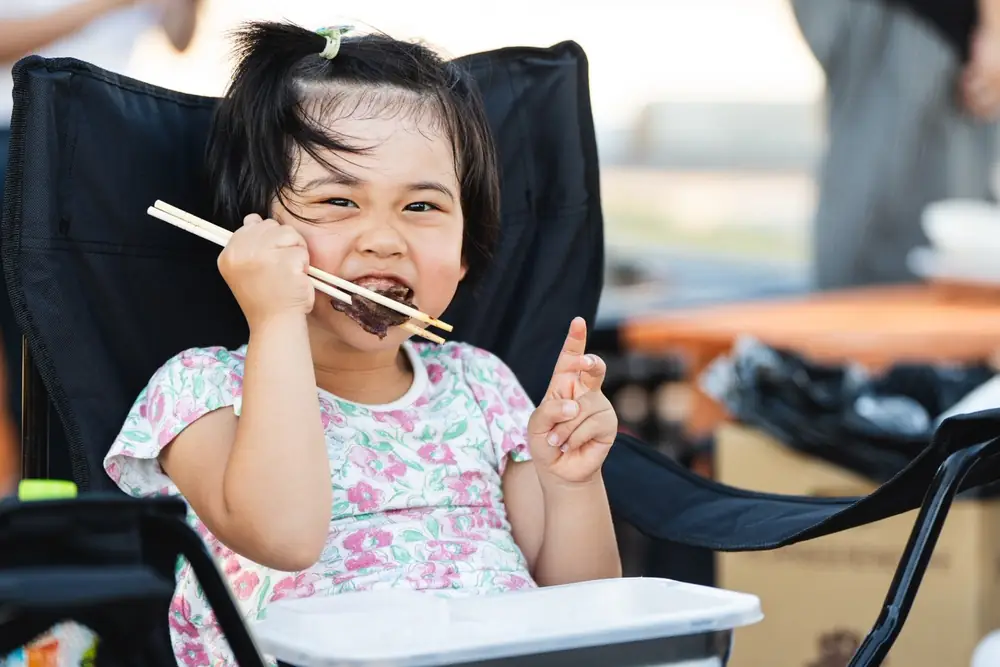アウトドアチェアに座った子どもが、箸でつまんだ焼き肉を頬張っている。