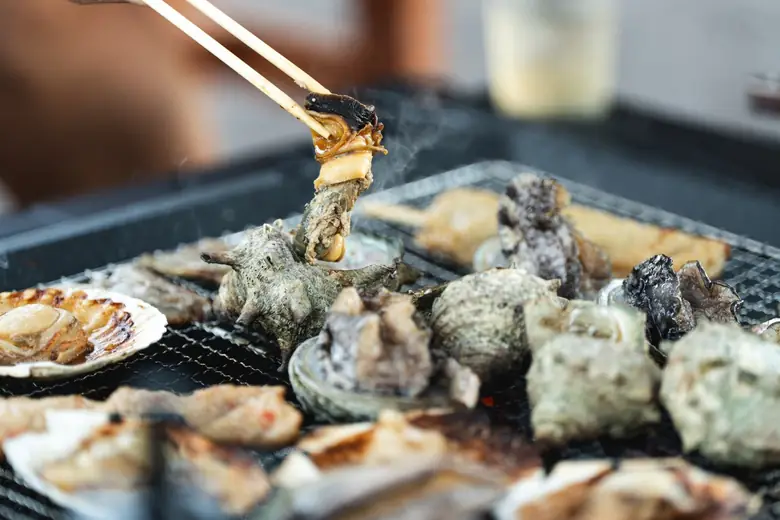 炭火の網の上でサザエやホタテを焼いているところ。箸でサザエの身を持ち上げている。